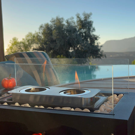 A modern outdoor fire pit table with a clear glass wind guard and a small flame, set on a wooden deck with an infinity pool and mountains in the background. A striped lounge chair and bowls of popcorn are partially visible.