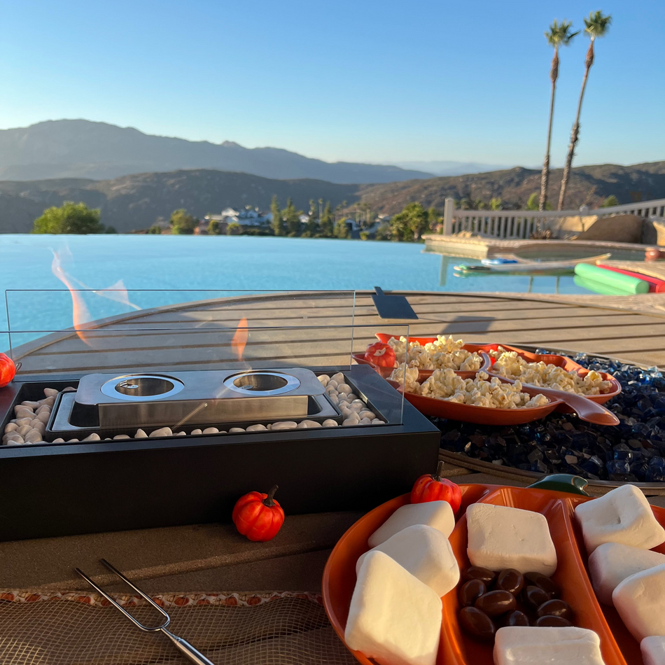 Outdoor patio scene with a black tabletop fire pit and flames, alongside marshmallows, popcorn, and a metal roasting fork, set beside an infinity pool with mountains and palm trees in the background.