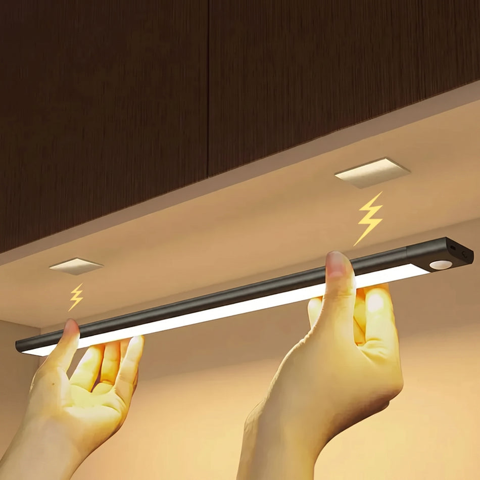 A person's hands are shown holding a slim black LED under-cabinet light bar, illuminating a kitchen cabinet space. The light bar is positioned horizontally beneath dark brown wooden cabinets, with yellow lightning bolt graphics indicating illumination.