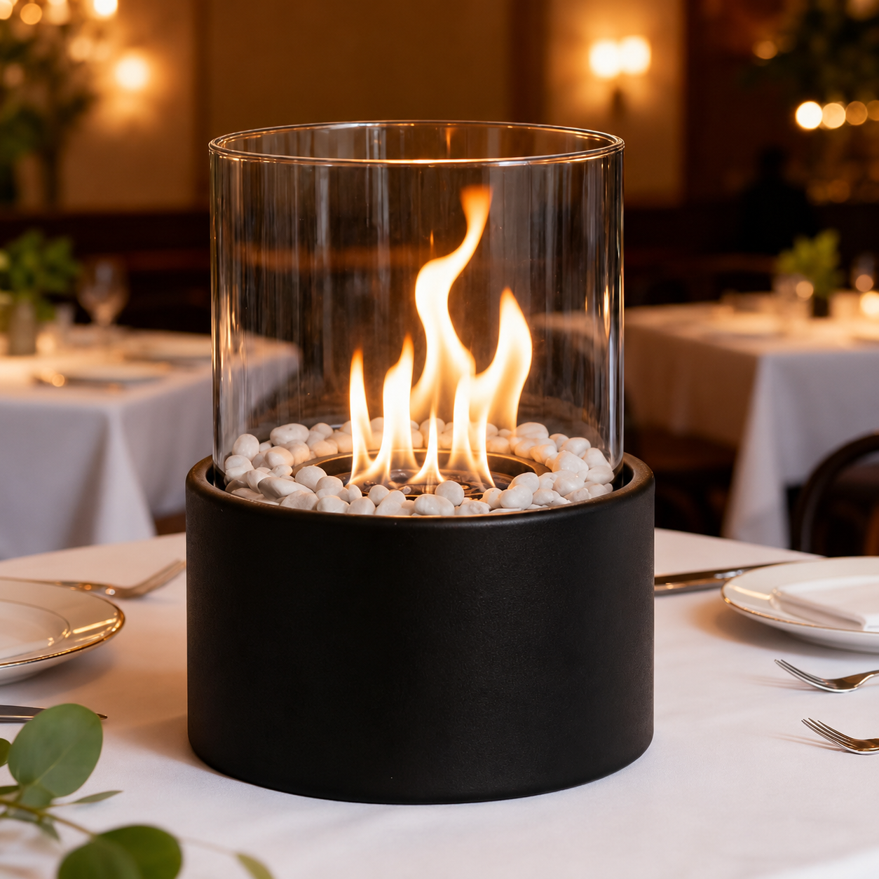 A modern tabletop fire pit with a matte black cylindrical base, white decorative stones, and a clear glass wind guard showing a warm orange flame, set on a white tablecloth in a softly blurred restaurant setting.