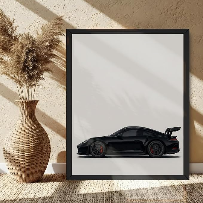 A framed art print featuring a black sports car in profile, displayed on a woven rug next to a wicker vase with dried pampas grass against a textured beige wall.