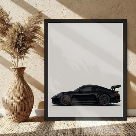 A framed art print featuring a black sports car in profile, displayed on a woven rug next to a wicker vase with dried pampas grass against a textured beige wall.