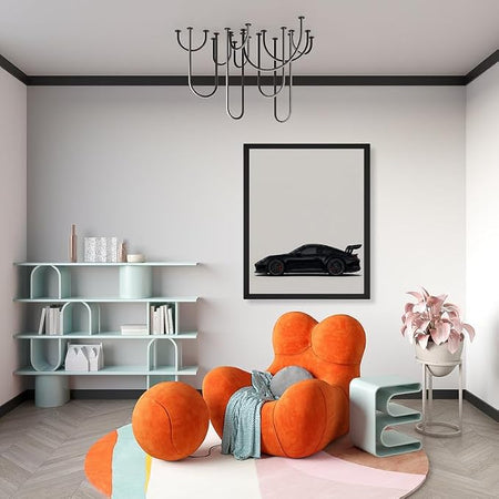 A modern living room corner featuring a bold orange cloud-shaped armchair with a blue throw blanket, a light blue shelving unit, a black framed poster of a sports car, and a pink plant on a white stand.